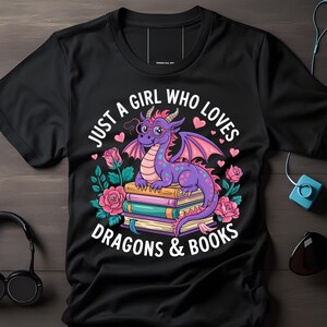 Può includere: T-shirt nero con un drago viola con gli occhiali, seduto sui libri. Il testo dice "JUST A GIRL WHO LOVES DRAGONS & BOOKS" circondato da rose rosa e cuori. Un design divertente.