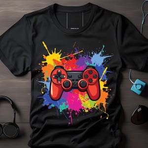 Puede incluir: Camiseta negra con un diseño de mando de videojuegos rojo y negro. El mando se encuentra sobre un vibrante fondo de salpicaduras de pintura coloridas. También se ven auriculares, gafas de sol y auriculares.
