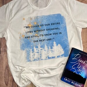 May include: White t-shirt with a blue and white watercolor design featuring a quote from the book "Beneath the Stars" by Emily McIntire. The quote reads: "We could go our entire lives without speaking, and still, I'd know you in the next one."
