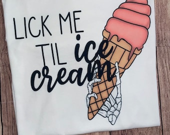 May include: White t-shirt with black text that reads "Lick Me Til Ice Cream" and a graphic of a skeleton hand holding a pink ice cream cone.
