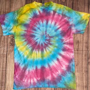 Może przedstawiać: Koszulka tie-dye z wirującym wzorem w odcieniach różowego, niebieskiego, żółtego i zielonego.