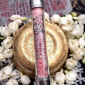 Puede incluir: Un colgante cilíndrico con tapas plateadas, que contiene un pergamino rosa y blanco con escritura negra y roja. Descansa sobre una esfera dorada grabada, rodeada de flores blancas. La URL de la tienda Etsy es visible.