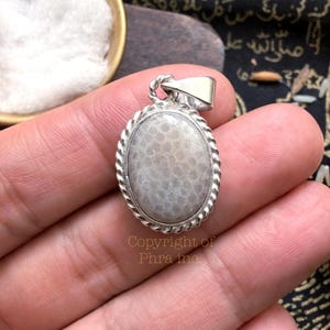 May include: An oval pendant featuring a smooth, light gray fossilized coral stone set in a polished silver frame with a twisted rope border design. The pendant includes a silver bail at the top and displays the text Copyright of Phra inc.
