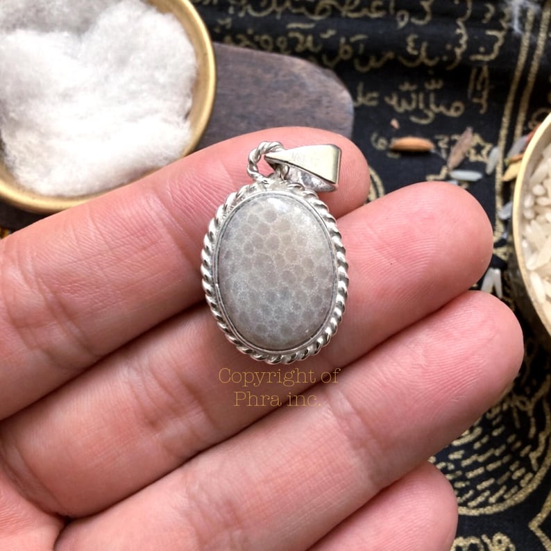 May include: An oval pendant featuring a smooth, light gray fossilized coral stone set in a polished silver frame with a twisted rope border design. The pendant includes a silver bail at the top and displays the text Copyright of Phra inc.