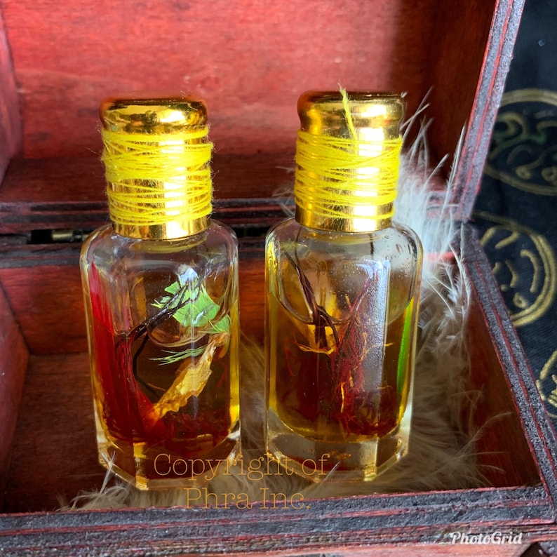 Puede incluir: Dos peque&ntilde;as botellas de vidrio transparente con tapas doradas envueltas en hilo amarillo. Cada botella contiene un l&iacute;quido con elementos bot&aacute;nicos rojos y verdes. Las botellas est&aacute;n dentro de una caja de madera con acabado rojo. El texto "Copyright of Phra Inc." es visible.