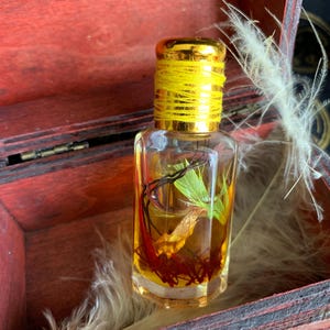 Puede incluir: Una peque&ntilde;a botella de vidrio transparente con una tapa dorada y un hilo amarillo alrededor del cuello. La botella contiene un l&iacute;quido dorado con hierbas y especias secas. La botella est&aacute; dentro de una caja de madera roja.