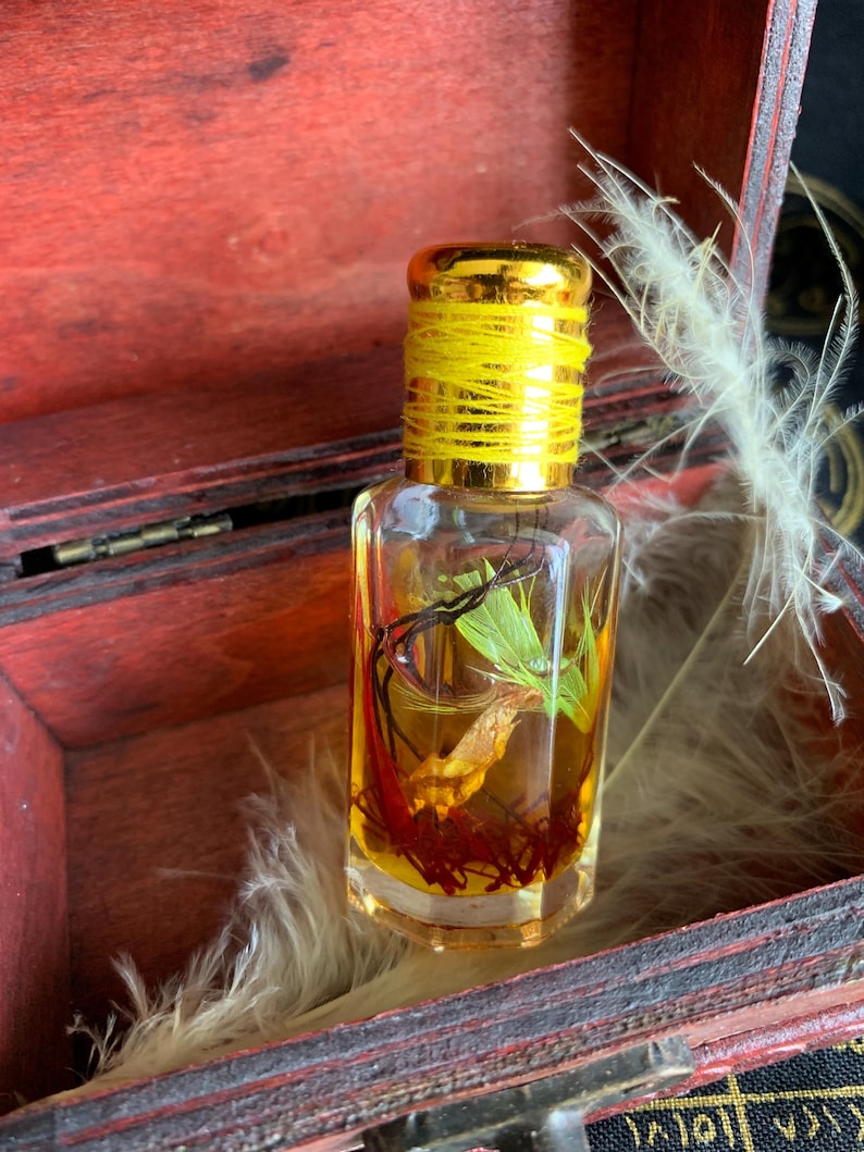 Puede incluir: Una peque&ntilde;a botella de vidrio transparente con una tapa dorada y un hilo amarillo alrededor del cuello. La botella contiene un l&iacute;quido dorado con hierbas y especias secas. La botella est&aacute; dentro de una caja de madera roja.