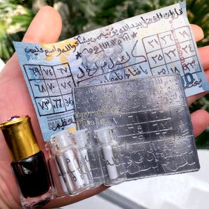 Op de afbeelding: Een klein glazen flesje met een gouden dop gevuld met donkere vloeistof, drie piepkleine doorzichtige glazen flesjes met opgerolde papieren, een rechthoekige metalen plaat met Arabisch schrift en een cijferrooster, en blauw papier.