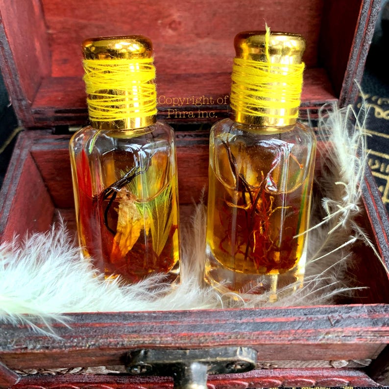 May include: Two small glass bottles with gold lids and yellow string detail. The bottles contain a golden liquid and botanical elements. They are presented in a wooden box with white feathers.
