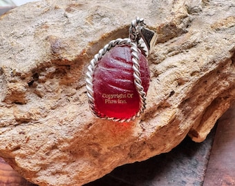 May include: A pendant featuring a translucent red scarab-shaped stone encased in a twisted silver-toned metal wire frame with a triangular bail. The stone surface displays horizontal carved lines and the text Copyright Of Phra Inc.