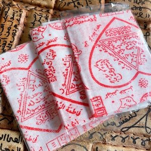 Puede incluir: Tres cuadrados de tela blanca con marcas de tinta roja. Las marcas están dispuestas en un patrón geométrico e incluyen escritura árabe. Los cuadrados de tela están doblados y empaquetados en una bolsa de plástico transparente.