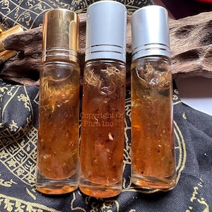May include: Three amber glass bottles with gold and silver roll-on tops filled with a clear liquid containing small, gold flecks and brown fibers. The bottles are sitting on a black fabric with gold designs.