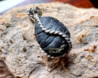 May include: A black, oval-shaped pendant with a textured surface, wrapped in a silver-colored twisted rope design. The pendant is set against a natural stone background. The text "Copyright Of Phra Inc." is visible.