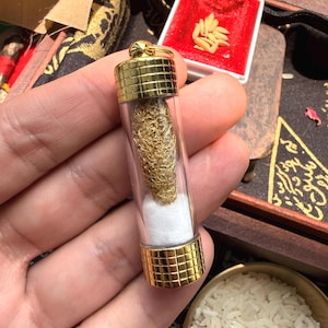 May include: A clear glass vial with gold-colored metal caps at each end, containing a gold-colored, woven object and white cotton. The vial is held in a hand, with a small red box and rice in the background.