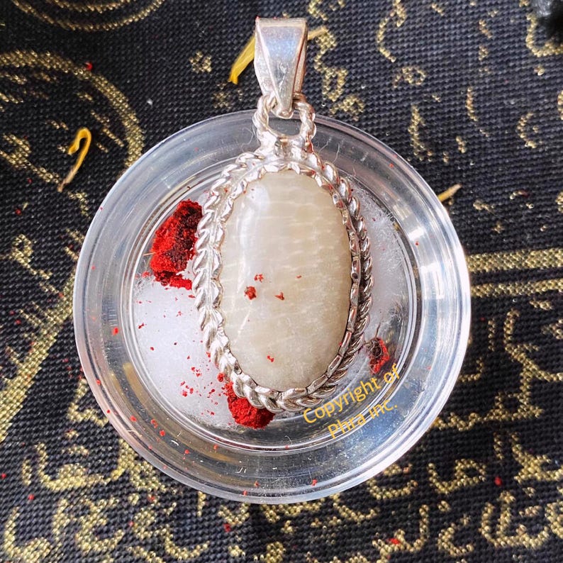 May include: An oval-shaped pendant featuring a smooth, off-white stone set in a twisted silver-toned metal frame with a bail at the top. The pendant rests inside a clear circular container with red powder and white granules. Text reads Copyright of Phra Inc.