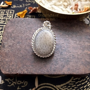 May include: An oval pendant featuring a smooth, light gray stone with a subtle honeycomb-like texture, encased in a twisted silver-toned metal border. The pendant includes a small, decorative silver-toned bail at the top for attachment.