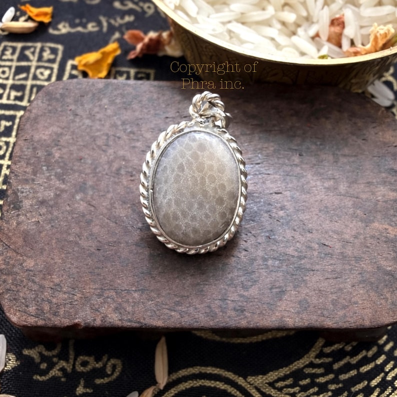 May include: An oval pendant featuring a smooth, light gray stone with a subtle honeycomb-like texture, encased in a twisted silver-toned metal border. The pendant includes a small, decorative silver-toned bail at the top for attachment.