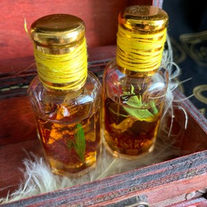 Puede incluir: Dos peque&ntilde;as botellas de vidrio transparente con tapas doradas y cuerda amarilla alrededor del cuello. Las botellas contienen un l&iacute;quido dorado con materia vegetal visible. Se exhiben en una peque&ntilde;a caja de madera abierta.