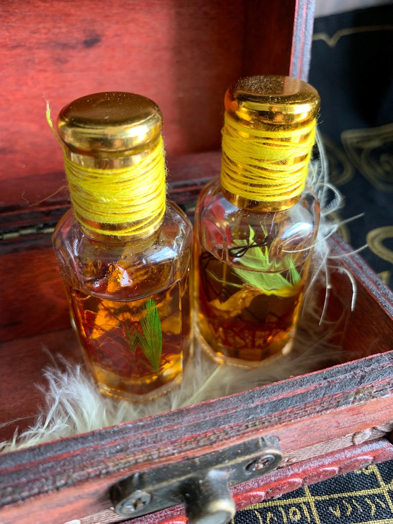 Puede incluir: Dos peque&ntilde;as botellas de vidrio transparente con tapas doradas y cuerda amarilla alrededor del cuello. Las botellas contienen un l&iacute;quido dorado con materia vegetal visible. Se exhiben en una peque&ntilde;a caja de madera abierta.