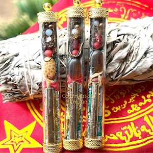 Op de afbeelding: Drie kleine glazen flesjes met gouden dopjes, elk gevuld met verschillende charmes en amuletten. De flesjes zijn geplaatst tegen een rode achtergrond met een wit en gouden ontwerp.