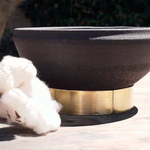 Könnte beinhalten: Eine mattschwarze Schale mit strukturierter Außenfläche, die an der Basis ein breites, glattes, goldfarbenes Metallband aufweist. Die Schale steht neben einem Büschel weißer, flauschiger Baumwollkapseln, was einen Kontrast zwischen dem dunklen Gefäß und der leichten, organischen Textur der Baumwolle erzeugt.
