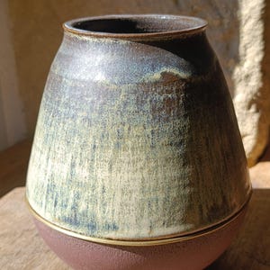 Peut inclure: Un vase en céramique à la silhouette arrondie, doté d'une partie supérieure émaillée lisse de couleur brun foncé et crème, qui se transforme en une base mate et texturée de couleur terre cuite, séparée par un fin rebord doré horizontal.