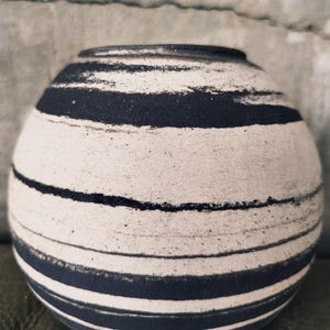 Peut inclure: Un vase en céramique sphérique avec une surface mate texturée blanc cassé, orné de rayures horizontales noires d'épaisseurs variables. Le récipient présente une petite ouverture circulaire au sommet et un corps arrondi qui se rétrécit vers la base.