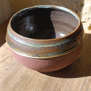 Op de afbeelding: Een ronde keramische kom met een textuurrijke, matte terracotta-kleurige buitenkant en een gladde, glanzend donkerbruine binnenkant. De bovenrand is afgewerkt met een decoratieve band van goudkleurig glazuur en subtiele blauwgroene accenten.