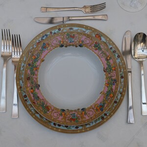 Puede incluir: Un plato de cena de porcelana blanca con un patr&oacute;n floral dorado y rosa. El plato est&aacute; colocado sobre un mantel blanco con un tenedor, cuchillo y cuchara de plata.