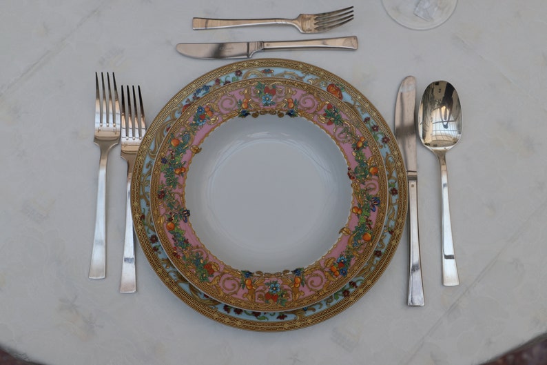 Puede incluir: Un plato de cena de porcelana blanca con un patr&oacute;n floral dorado y rosa. El plato est&aacute; colocado sobre un mantel blanco con un tenedor, cuchillo y cuchara de plata.
