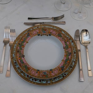Puede incluir: Un plato de porcelana blanca con un patr&oacute;n floral colorido y detalles dorados. El plato est&aacute; puesto con un tenedor, un cuchillo y una cuchara.