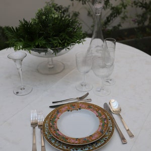 Puede incluir: Una mesa puesta con un mantel blanco, un plato decorativo con un dise&ntilde;o floral dorado y rosa, cubiertos de plata y copas de vino de vidrio transparente.