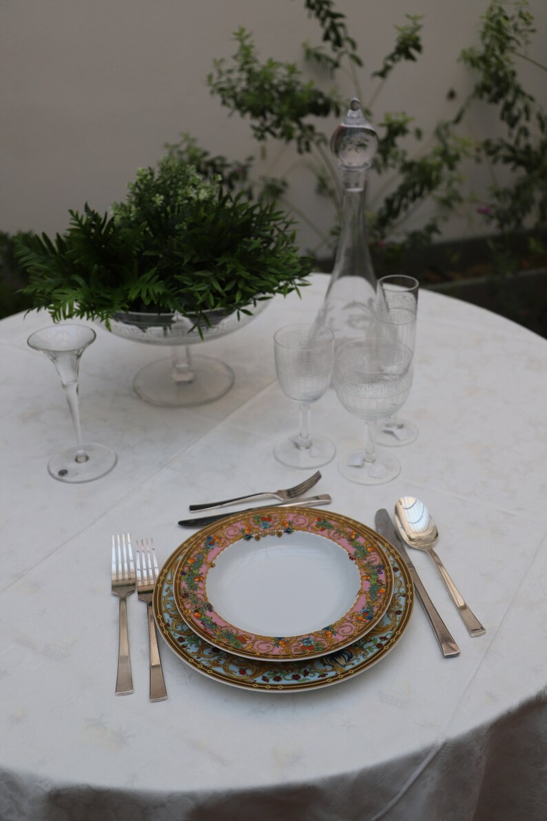 Puede incluir: Una mesa puesta con un mantel blanco, un plato decorativo con un dise&ntilde;o floral dorado y rosa, cubiertos de plata y copas de vino de vidrio transparente.