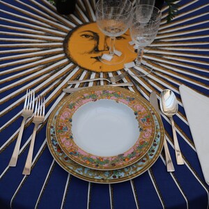 Puede incluir: Un plato blanco para cenar con un estampado floral colorido se encuentra sobre un mantel a rayas azul y dorado. Dos tenedores de plata, un cuchillo de plata y una cuchara de plata se colocan junto al plato. Una servilleta blanca est&aacute; doblada a la derecha del plato.