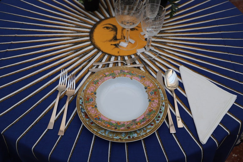 Puede incluir: Un plato blanco para cenar con un estampado floral colorido se encuentra sobre un mantel a rayas azul y dorado. Dos tenedores de plata, un cuchillo de plata y una cuchara de plata se colocan junto al plato. Una servilleta blanca est&aacute; doblada a la derecha del plato.