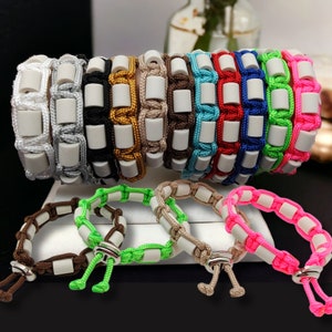 Könnte beinhalten: Ein Set aus 12 bunten Armbändern mit weißen Perlen. Jedes Armband besteht aus geflochtener Schnur und hat eine einzigartige Farbkombination. Die Armbänder sind in einer Reihe angeordnet, wobei die Farben von hell nach dunkel übergehen.