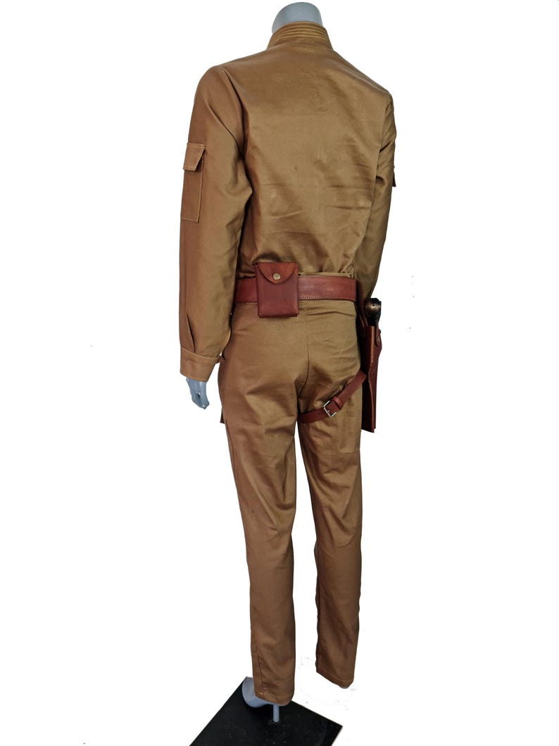May include: A tan jumpsuit with a brown leather belt and a holster. The jumpsuit has a collared shirt with long sleeves and a zipper closure. The pants have a straight leg and a zipper fly.