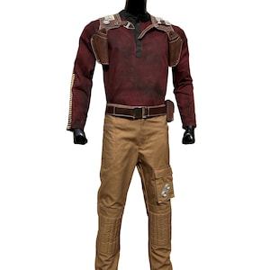 Può includere: Una camicia a maniche lunghe marrone e rossa con un gilet in pelle marrone e una cintura marrone. La camicia ha un aspetto consumato e il gilet ha tasche e cinturini. La persona indossa pantaloni marroni con tasche cargo e ginocchiere.