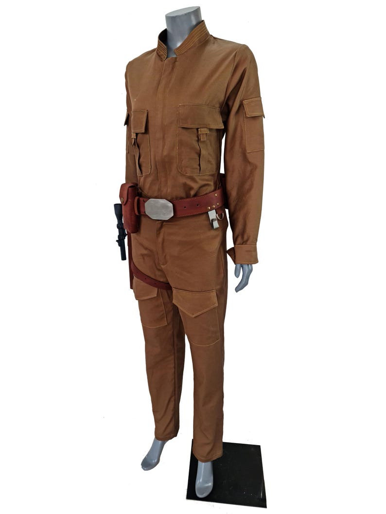 May include: A brown jumpsuit with a brown belt and a silver buckle. The jumpsuit has a collared shirt with a mandarin collar and long sleeves. The jumpsuit has two pockets on the chest and two pockets on the legs. The jumpsuit is worn by a mannequin.