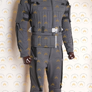 May include: A grey tactical suit with a quilted texture, featuring a high collar, a front zip, and multiple pockets. The suit includes a belt with a white buckle and black straps. The mannequin is wearing a matching grey helmet.