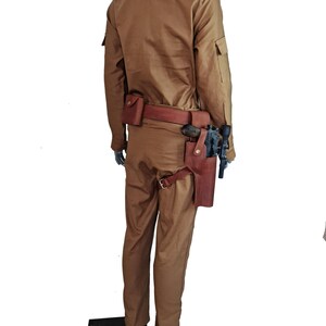 May include: A tan jumpsuit with a brown leather belt and a holster with a gun. The jumpsuit has a zipper and buttons on the front.
