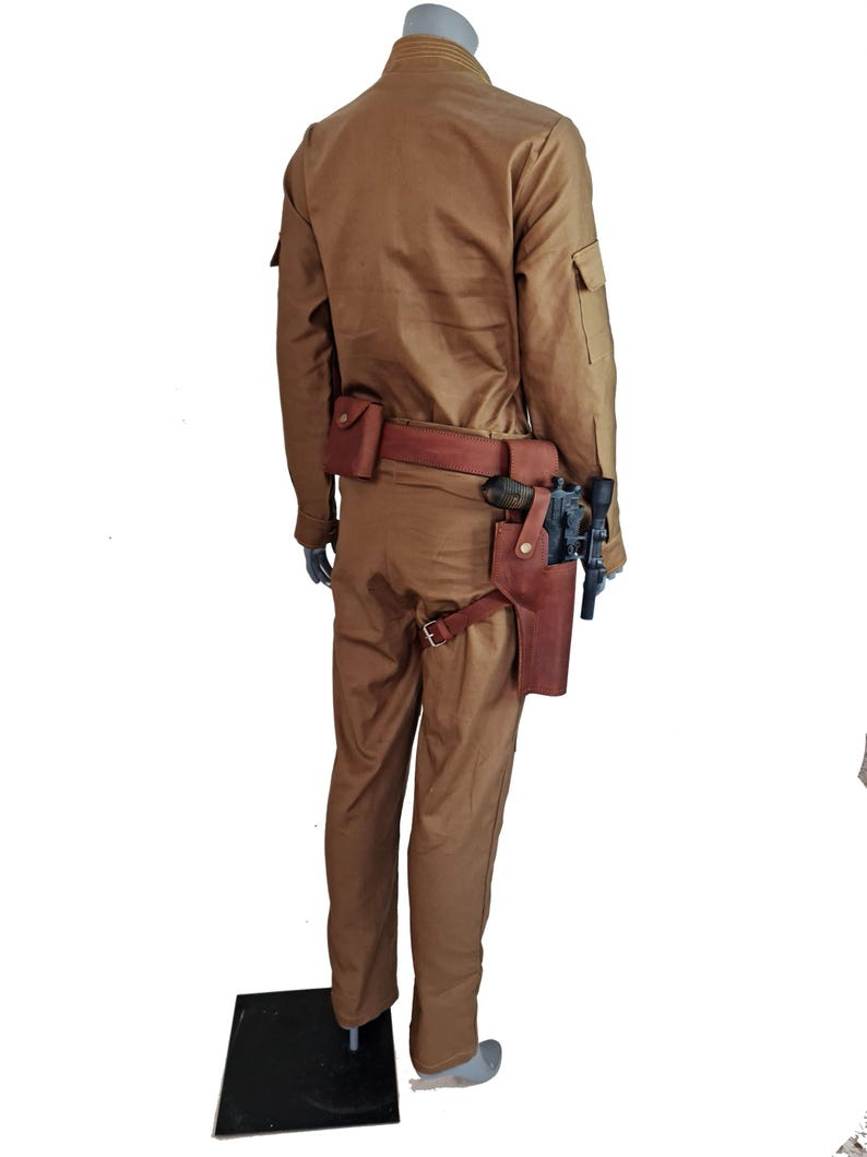 May include: A tan jumpsuit with a brown leather belt and a holster with a gun. The jumpsuit has a zipper and buttons on the front.