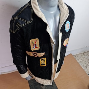 May include: A black quilted jacket with a white collar and a white lining. The jacket has several patches sewn on the front, including a yellow and black patch with a symbol, a gold patch with wings, and a red, white, and blue patch with a symbol.