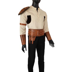 May include: A white long-sleeved shirt with brown leather shoulder pads and matching arm cuffs. A brown leather belt with a metal buckle is worn over the shirt. Black pants complete the outfit.