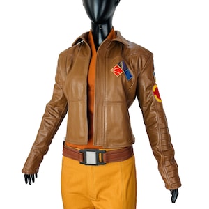 Könnte beinhalten: Braune Lederjacke mit passendem Gürtel und gelber Hose. Die Jacke hat Aufnäher auf den Schultern und Ärmeln. Das Outfit wird auf einer schwarzen Schaufensterpuppe vor weißem Hintergrund präsentiert.
