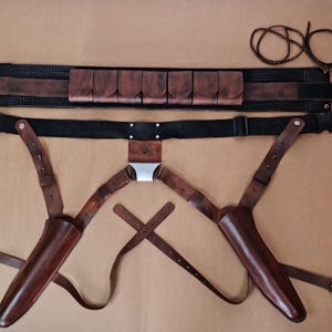 Op de afbeelding: Een bruine leren riem- en holsterset. De set bevat een riem met zakjes, een zwarte riem, twee holsters en een set riemen. Het leer heeft een rijke, donkerbruine kleur en het ontwerp lijkt handgemaakt.