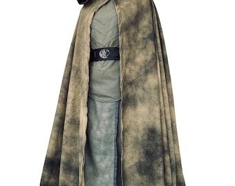 May include: A hooded cloak with a brown and tan camouflage pattern. The cloak is worn over a gray tunic and pants. A black belt with a circular buckle is worn around the waist.