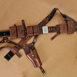 Könnte beinhalten: Ein brauner Ledergürtel mit mehreren Taschen und einem Holster. Der Gürtel hat eine silberne Schnalle mit einem Design. Das Holster ist mit Riemen am Gürtel befestigt und hat eine Klappe, die die Öffnung bedeckt.