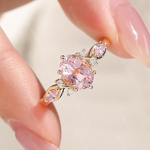 May include: A gold ring featuring a central oval-cut pink gemstone flanked by two smaller marquise-cut pink stones on each side, accented with tiny clear round stones set along the band in a delicate, symmetrical arrangement.