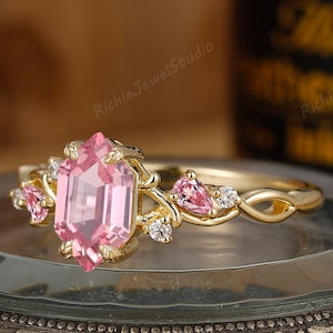 May include: A gold ring with a large, pink, emerald-cut gemstone as the centerpiece. The ring features smaller pink gemstones and small white stones. The band has a delicate, intertwined design. The ring is displayed on a vintage-style, brass-colored box.
